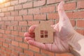 Close-up of a small wooden house in hand against a brick wall background. Concept - security, purchase, construction of real