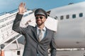 Cheerful aircraft pilot standing outdoors at airport and waving hello to colleagues