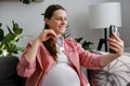 Close up of smiling beautiful young pregnant woman sitting on cozy couch holding ultrasound pictures of baby with family using