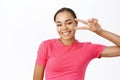 Close up of smiling black female model, shows peace, v-sign near eye and looks happy, wears pink tshirt, stands over