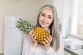 Close up of smiling elderly gray haired woman in white shirt, holding fresh pineapple while posing to camera on light