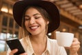 Close up of a smiling lovely girl in hat sitting at the cafe table indoors, holding cup of tea, Royalty Free Stock Photo