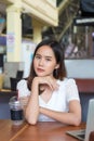 Close up smiling young Asian woman sitting in front of laptop on wooden table put her chin on her hand, looking at camera at Royalty Free Stock Photo