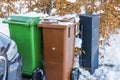 Close-up of snow covered trash bins for sorting garbage next to black mailbox against dry hedges background.