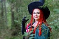 Close-up Sorceress with red hair in a hat and a black cloak in the woods in the hand with a candle.