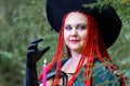 Close-up Sorceress with red hair in a hat and a black cloak in the woods in the hand with a candle.