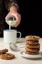 Close-up of stack of chocolate cookies on white plate and woman`s hand serving milk in white cup, selective focus, on white woode Royalty Free Stock Photo
