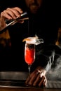 Close-up of stemware with smoked drink and hand of bartender pouring beverage from jigger into it