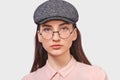 Close up studio portrait of pretty young woman wears transparent spectacles, casual pink shirt and trendy gray cap, looks Royalty Free Stock Photo