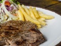 Close up of T-bone steak with salt and pepper, golden french fries and green vegetables. on white plate, Fork and knife, wooden ta Royalty Free Stock Photo