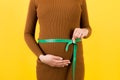 Close up of tape measure on pregnant woman`s belly in brown dress at yellow background. Measuring of growing abdomen. Copy space Royalty Free Stock Photo