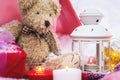 A close-up of a teddy bear with Christmas candles on a cozy checkered plaid outdoors next to a red gift box sprinkled