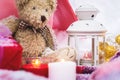 A close-up of a teddy bear with Christmas candles on a cozy checkered plaid outdoors next to a red gift box sprinkled