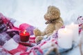 A close-up of a teddy bear with Christmas candles on a cozy checkered rug in the outdoors next to a snowdrift sprinkled