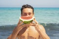 Close up of teenager holding watermelon in front of face. Portrait of shirtless teenage boy holding halved watermelon fruit and