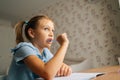 Close-up of thinking primary little child girl doing homework and holding pen against mouth sitting at home table by Royalty Free Stock Photo