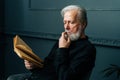 Close-up of thoughtful senior gray-haired aged male reading paper book sitting on chair in dark living room, relax and Royalty Free Stock Photo