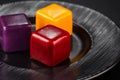 Close-up three miniature cakes in the shape of a cube of different colors on a black plate.
