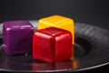 Close-up three miniature cakes in the shape of a cube of different colors on a black plate.