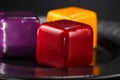 Close-up three miniature cakes in the shape of a cube of different colors on a black plate.