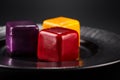 Close-up three miniature cakes in the shape of a cube of different colors on a black plate.