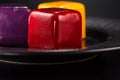 Close-up three miniature cakes in the shape of a cube of different colors on a black plate.