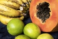 Close up to a fresh sliced orange papaya with seeds inside, a freckled bananas cluster and three green lemons over a blue navy bac Royalty Free Stock Photo