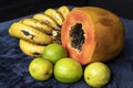 Close up to a fresh sliced orange papaya with seeds inside, a freckled bananas cluster and three green lemons over a blue navy and Royalty Free Stock Photo