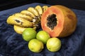 Close up to a fresh sliced orange papaya with seeds inside, a freckled bananas cluster and three green lemons over a blue navy and Royalty Free Stock Photo