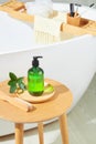 Close-up of a transparent green shampoo bottle placed on a ceramic plate, green leaves and a brush on a wooden table. Behind is