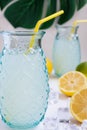Close-up of two glasses with lemon juice with straws, on white wooden table with half lemons, lime and ice, selective focus, on wh