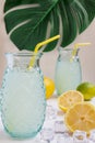 Close-up of two glasses with lemon juice with straws, on white wooden table with half lemons, lime and ice, on white background wi