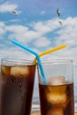 Close-up of two glasses of soda with ice and straws on the beach with blue sky and seagulls flying in Tenerife, Canary Islands