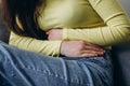 Close up of unknown sick young caucasian female suffering from stomach ache sitting on sofa at home, holding belly, feeling