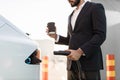 Close up of man holding coffee and charging electro car Royalty Free Stock Photo