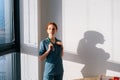 Close-up of unrecognizable female doctor in blue green uniform putting on stethoscope on neck standing near window in