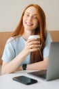 Close-up vertical portraitof happy young beautiful woman holding cup with hot coffee sitting at desk with laptop at cozy
