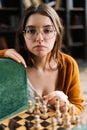 Close-up vertical portrait of young woman in elegant eyeglasses making chess move with knight piece sitting in floor Royalty Free Stock Photo