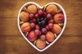 Close-up view from above of a white heart-shaped plate full of red grapes, apricots, strawberry and blueberries. Wooden background Royalty Free Stock Photo