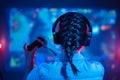 Close-up view from back of gamer girl playing video game at home in front of big screen with joystick and headphone. Colorful neon