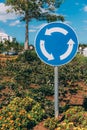 Close-up view of blue and white roundabout sign on the street with grass, trees, road and blue sky with clouds.