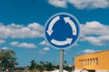 Close-up view of blue and white roundabout sign on the street with grass, trees, road and blue sky with clouds.