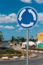 Close-up view of blue and white roundabout sign on the street with grass, trees, road and blue sky with clouds.