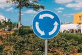 Close-up view of blue and white roundabout sign on the street with grass, trees, road and blue sky with clouds.