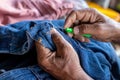 Close-up view of both hands of an elderly Thai man using a needle to pick up threads to repair jeans