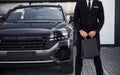 Close up view of businessman in black suit and tie stands near modern automobile with shopping bags in hands