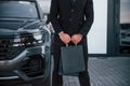 Close up view of businessman in black suit and tie stands near modern automobile with shopping bags in hands