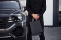 Close up view of businessman in black suit and tie stands near modern automobile with shopping bags in hands