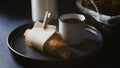Close up view of fresh baked croissant wrapped on piece of paper with a fresh hot coffee cup on a black plate simple morning