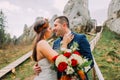 Close up view handsome groom in stylish blue suit kissing white dressed bride holding bouquet of roses on majestic